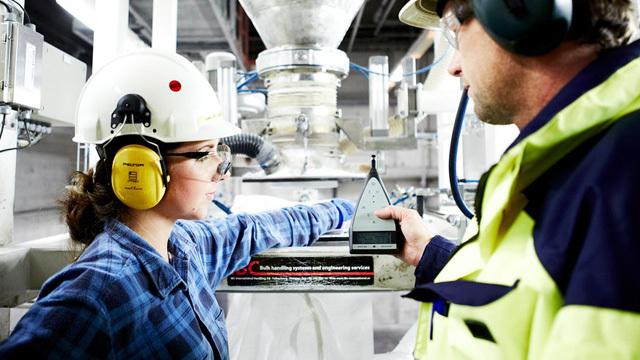 En kvinne med hjelm og hørselsvern viser en måler til en mann i arbeidsklær i en industriell setting.