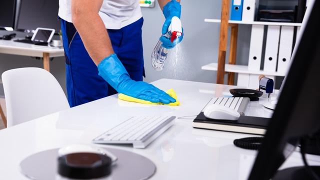 En person med blå hansker rengjør et skrivebord med en sprayflaske og en gul klut.