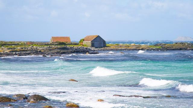 To hus med røde tak ved kysten, bølger som slår mot steinene og blått hav i bakgrunnen.