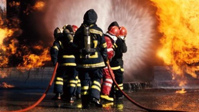 Brannmenn i uniformer slukker en stor brann med slanger, omringet av flammer og røyk.