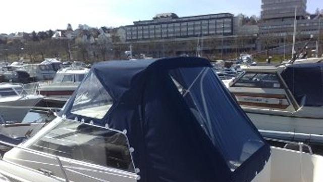 Båt med blåt kapell i marina, med flere båter og bygninger i bakgrunnen.