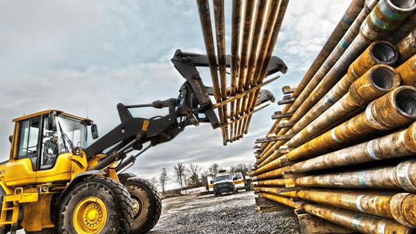 En gult gravemaskin løfter metallrør fra bakken og plasserer dem på en stabel av rør.