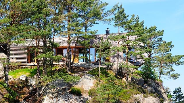 Moderne hytte i skogen, bygget av tre, med store vinduer og steinete landskap rundt.