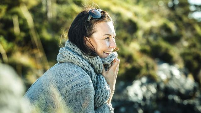 Kvinne med mørkt hår iført en grå genser og stor skjerf, smilende mens hun sitter ute i naturen.