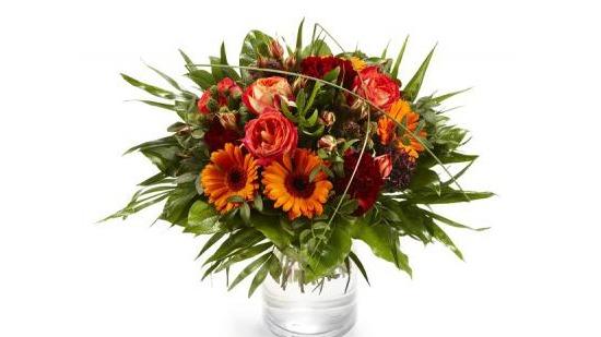 En fargerik blomsterbukett med røde roser, oransje gerberaer og grønt bladverk i en glassvase.