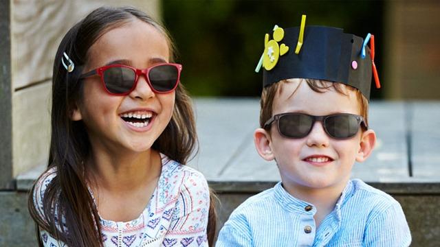 To barn med solbriller smiler mot kameraet, det ene barnet har en fargerik hatt på hodet.