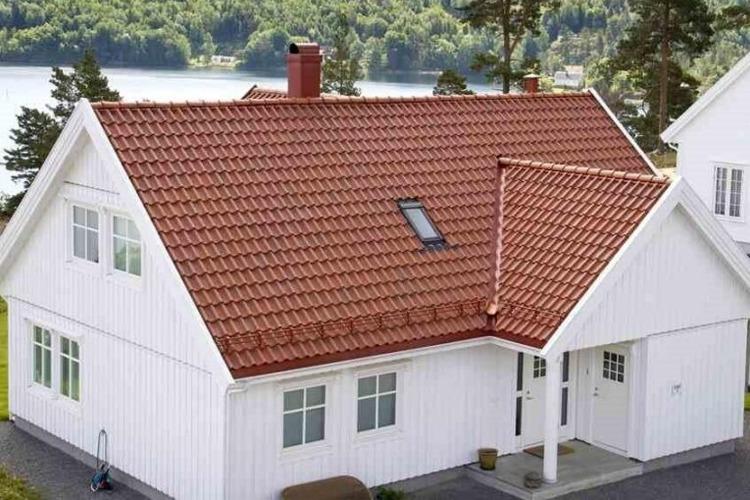 Moderne, hvitt hus med rød takstein, beliggende ved vannet med trær i bakgrunnen.