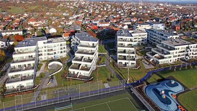 Moderne leilighetskompleks med flere bygninger, grøntområder og en tennisbane i bakgrunnen.