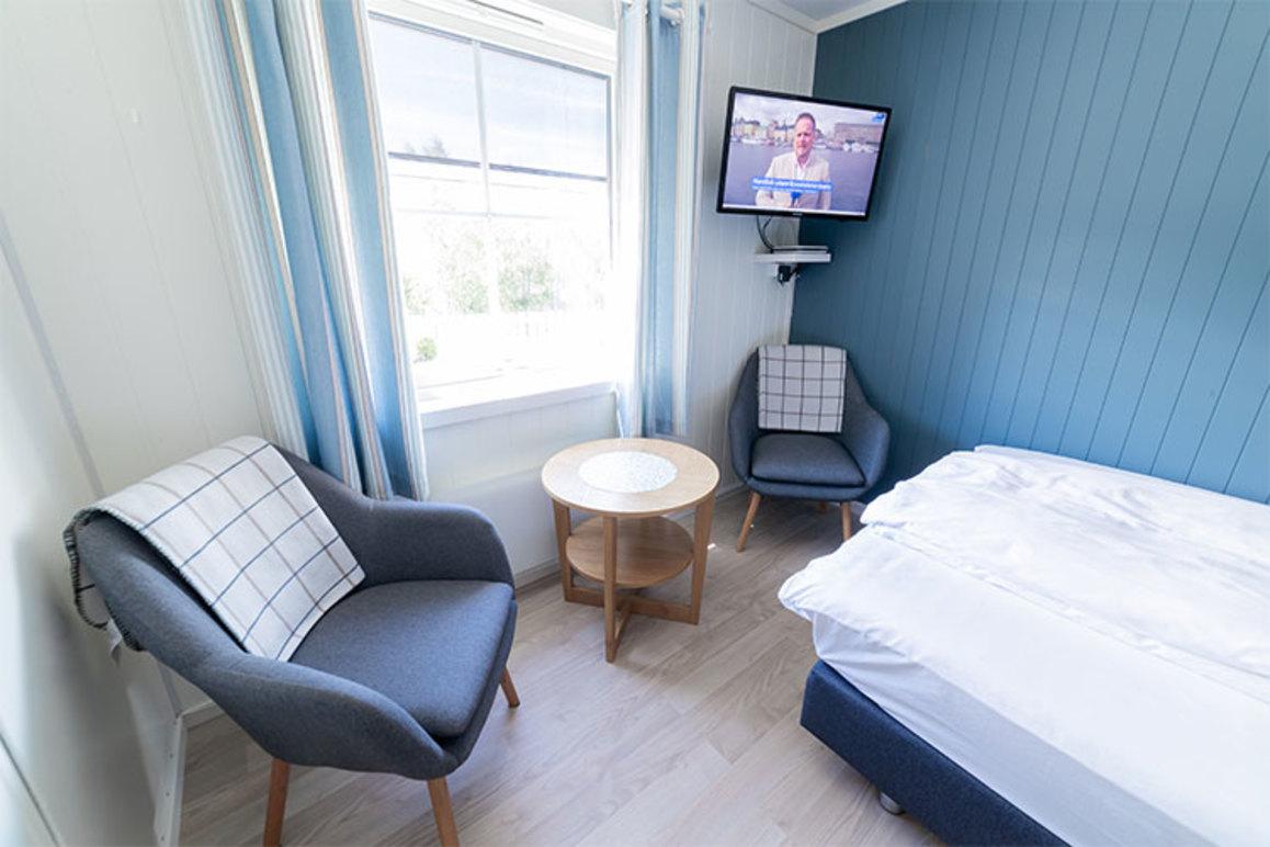 Koselig rom med to stoler, et bord, en seng og en TV på veggen. Naturlig lys strømmer inn fra vinduet.