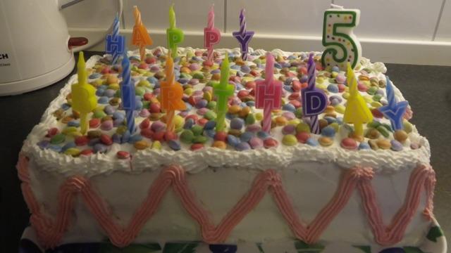 En bursdagskake med fargerike lys, pyntet med godteri og teksten "Happy 5th Birthday" på toppen.