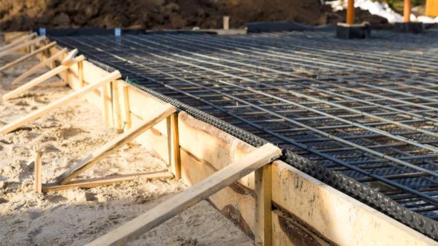 Bilde av en byggeplass med armeringsjern og forskaling for betongfundament. Sand og byggeutstyr er synlig.