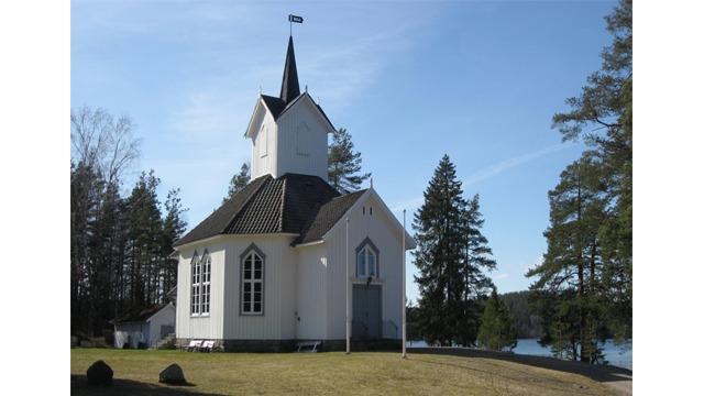 En hvit kirke med spisstårn, omringet av trær og en innsjø i bakgrunnen, under en klar blå himmel.