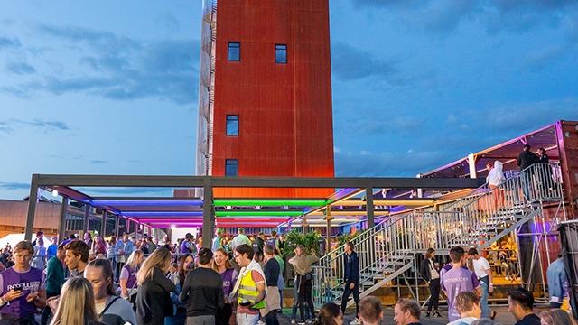 Folk samles på en takterrasse med fargerik belysning og en høy bygning i bakgrunnen under en kveldshimmel.