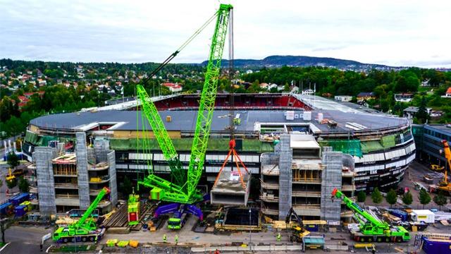Byggearbeid på stadion med store kraner og stillaser rundt bygningen. Grønn kran løfter betongelementer.