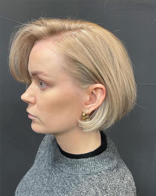 Kvinne med kort, blond hårfrisyre og gulløredobber, iført en grå genser, sett fra siden.