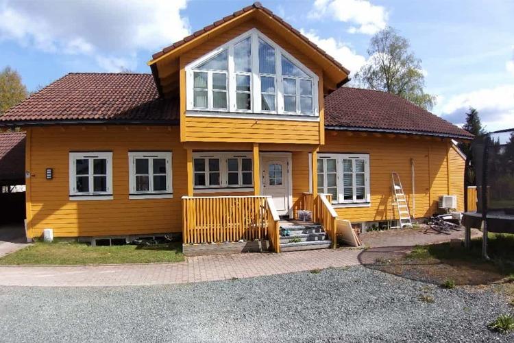 Gult hus med stor veranda og mange vinduer, omgitt av grønt landskap og blå himmel.