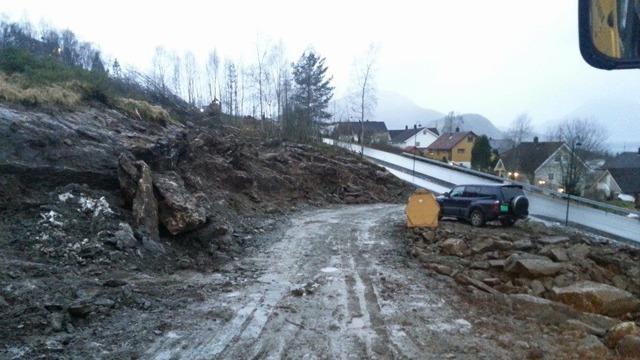 En vei med mudder og steiner etter et ras, med en bil parkert i nærheten av husene i bakgrunnen.