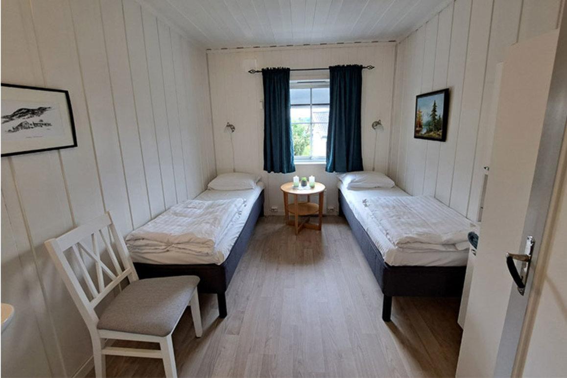 Rom med to enkeltsenger, et lite bord og stoler, samt vindu med gardiner. Veggene er hvite og gulvet er lyst.