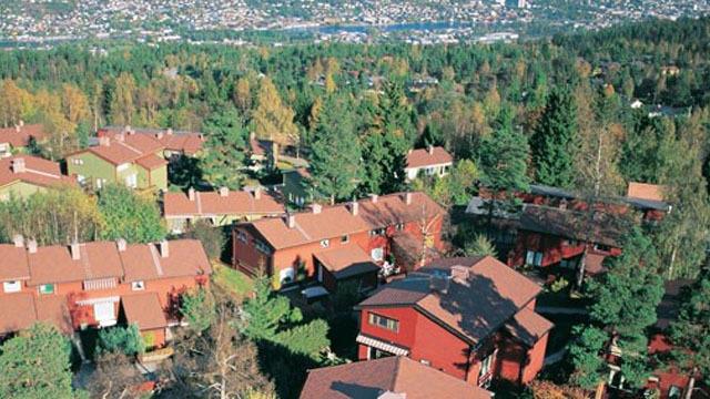Utsikt over et boligområde med røde hus omgitt av trær og grønt landskap. Fjell og by i bakgrunnen.