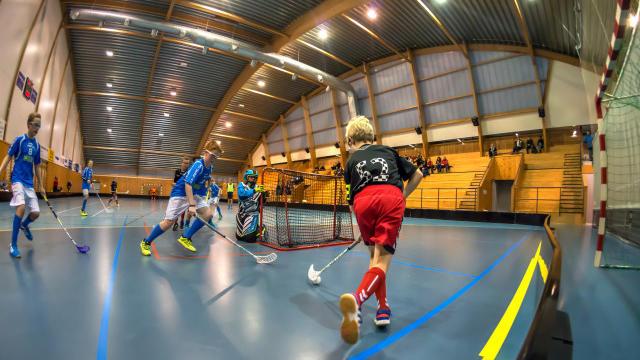 Ungdommer spiller innebandy i en idrettshall, med fokus på en spiller i svart drakt som løper mot målet.
