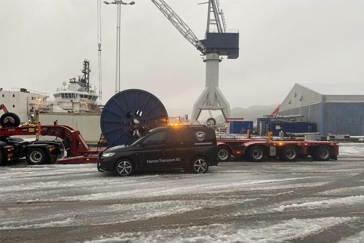 En lastebil med en stor kabeltrommel står på en snødekt havn, med kraner og skip i bakgrunnen.
