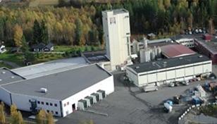 Oversiktsbilde av en fabrikk med flere bygninger og lagringsområder omgitt av trær og grønt landskap.