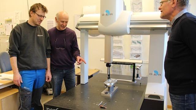Tre menn står ved en stor målemaskin i et verksted, og diskuterer måleprosessen.