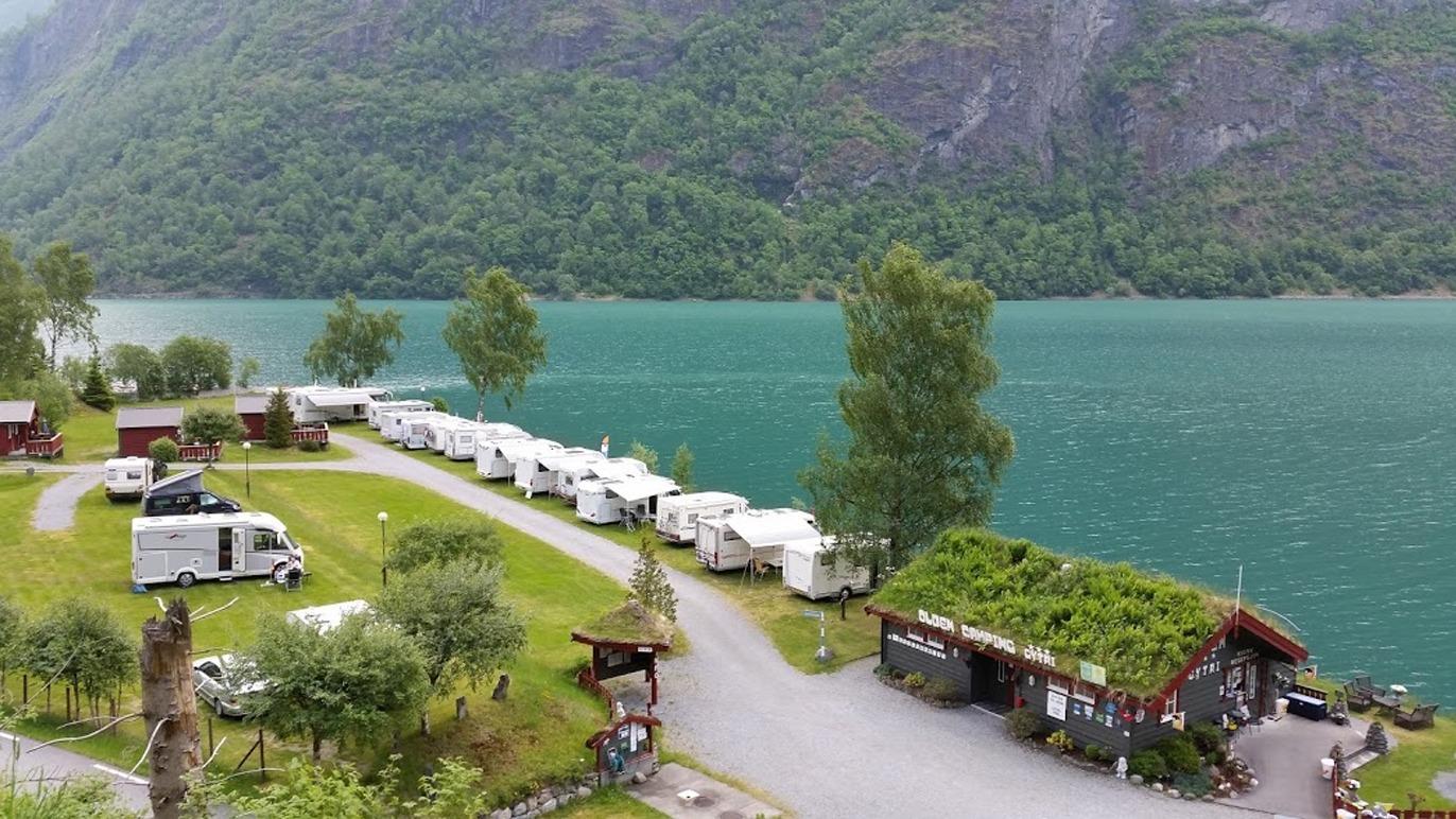 Campingplass ved en innsjø med campingvogner og en hytte omgitt av fjell og trær.