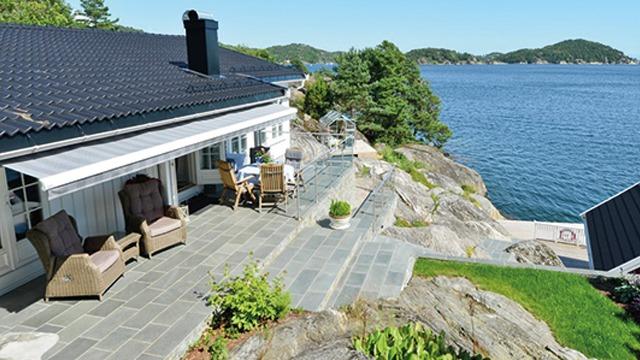Utsikt over en moderne hytte med terrasse, steinbelagt område og sjøutsikt mot grønne øyer.