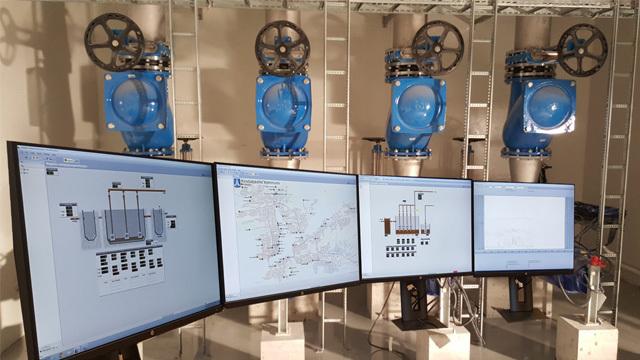 Moderne industriutstyr med blå ventiler og tre skjermer som viser tekniske diagrammer og kontroller.