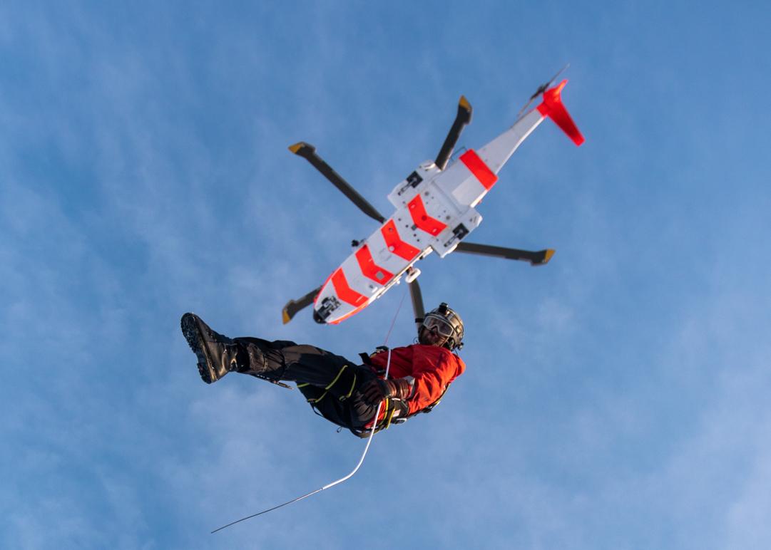 En redningsmann svever ned fra en helikopter med rød og hvit farge mot en blå himmel.