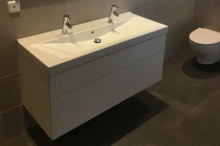 Moderne baderomsmøbler med en hvit servant og to kraner, plassert på en grå flislagt vegg.