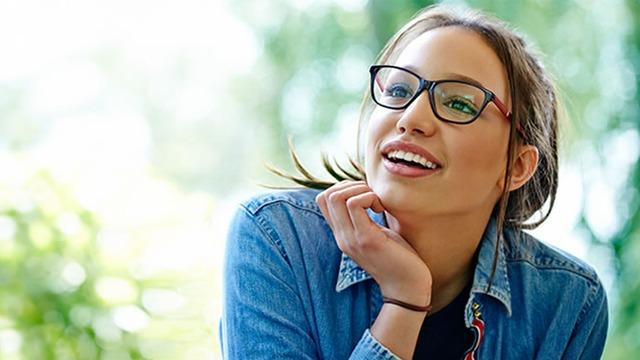 Ung kvinne med briller smiler og ser tankefull ut, iført en denimjakke i en grønn hage.