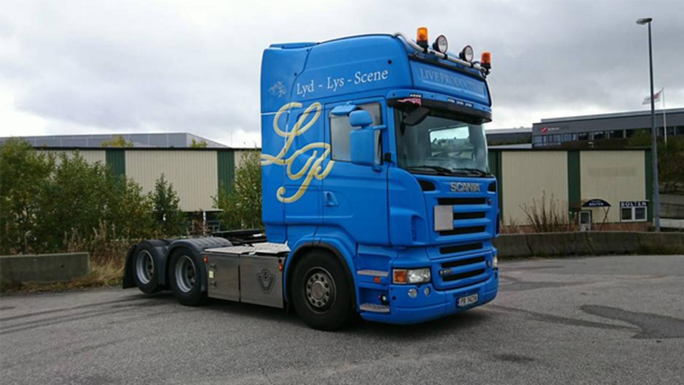 Blå lastebil med tre aksler parkert på en industriområde, med lys og dekorative detaljer.