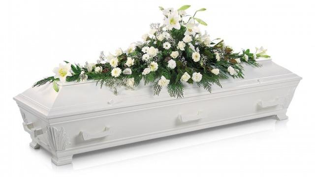 Hvit kiste dekorert med hvite blomster og grønt, plassert på en lys bakgrunn.