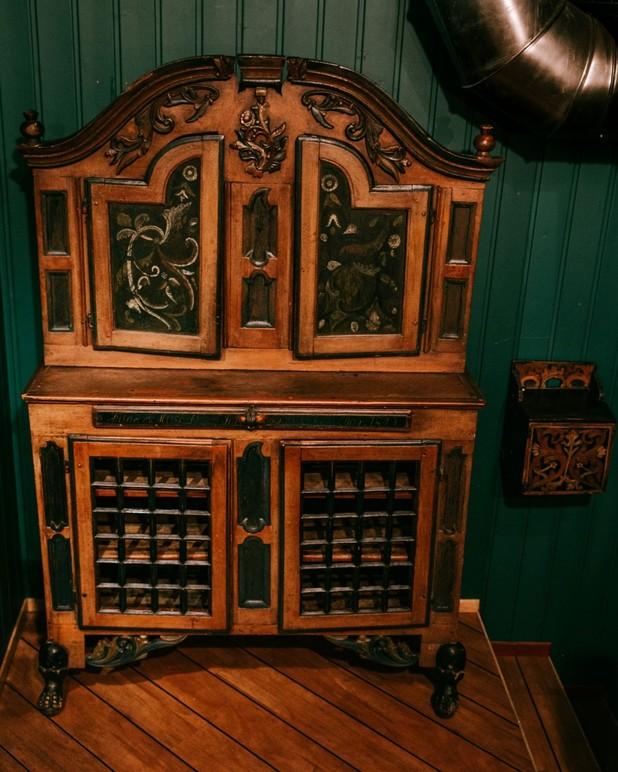 Et gammelt, dekorativt skap med detaljerte utskjæringer og glassdører, plassert mot en grønn vegg.