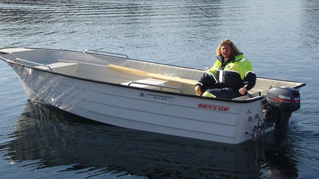 En person sitter i en liten båt på vannet, iført en gul flytedress og med motoren i bakgrunnen.