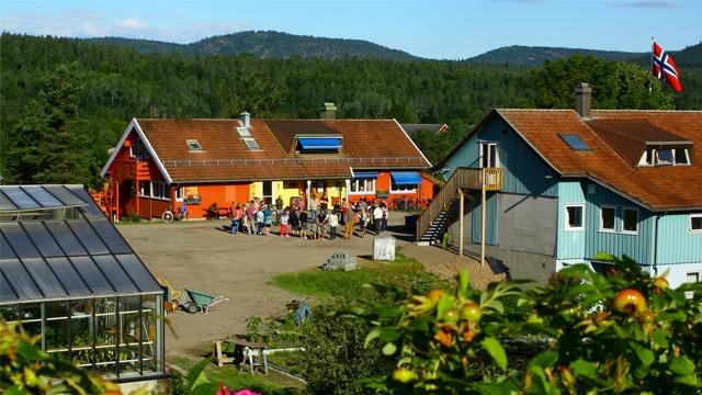 En fargerik gård med flere bygninger, folk samlet ute, og fjell i bakgrunnen. Solfylt dag med blå himmel.