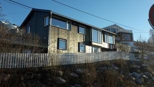 Moderne hus med store vinduer og trepanel, omgitt av en hvit gjerde og steiner i solfylt vær.