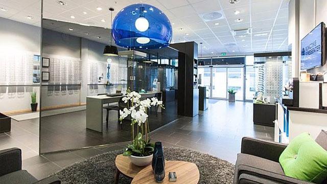 Moderne kontorinnredning med blå lampe, planter og sitteplasser i et lyst rom med glassvegger.