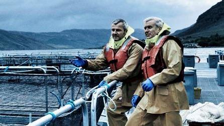 To menn i gule arbeidsdrakter og redningsvester står ved en fiskeoppdrettsanlegg med sjøen i bakgrunnen.