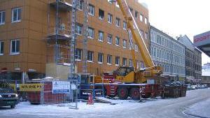 Byggeplass med kran og stillas, snødekt gate med bygninger i bakgrunnen.
