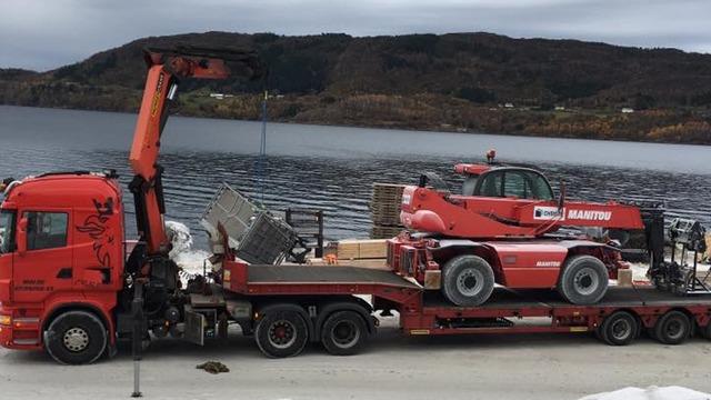 En lastebil med kran frakter en rød teleskoplaster på en tilhenger ved vannkanten.