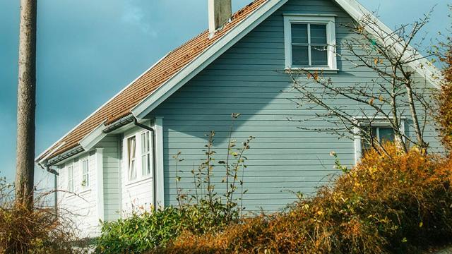 Blått hus med skråtak og hvite vinduer, omringet av busker og høstfarger.