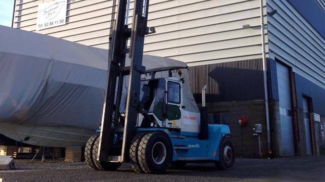 Gaffeltruck parkert ved en bygning, med en båt dekket av presenning i bakgrunnen.