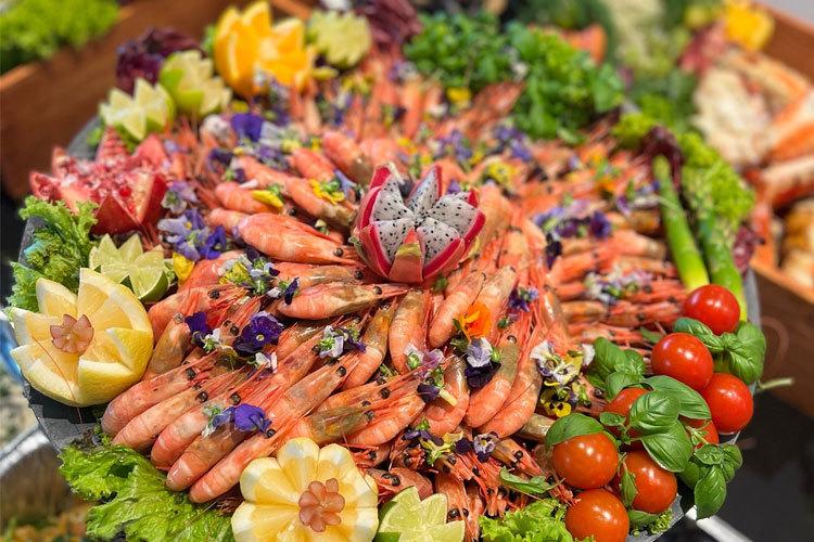 En fargerik sjømatfat med reker, grønnsaker, spiselige blomster og dekorative fruktskulpturer.