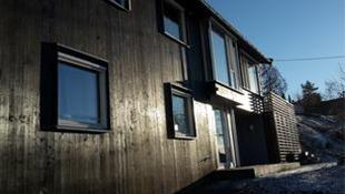 Moderne trehus med store vinduer, beliggende i solfylt område med snø på bakken.