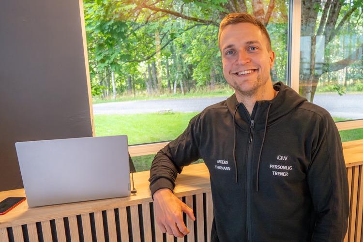 En smilende personlig trener står ved en laptop i et lyst rom med grønne trær i bakgrunnen.