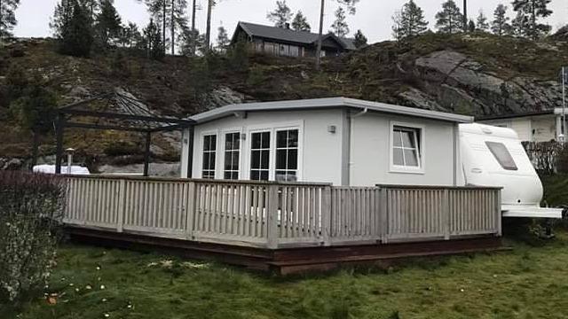 Moderne hytte med terrasse og campingvogn, omringet av natur og fjell i bakgrunnen.