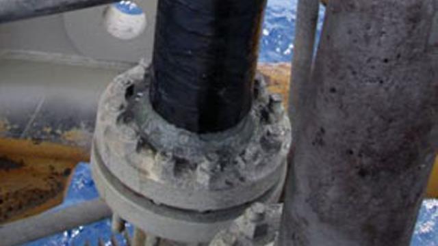 Nærbilde av en rørforbindelse på en offshore oljeplattform med synlig rust og sjøvann i bakgrunnen.